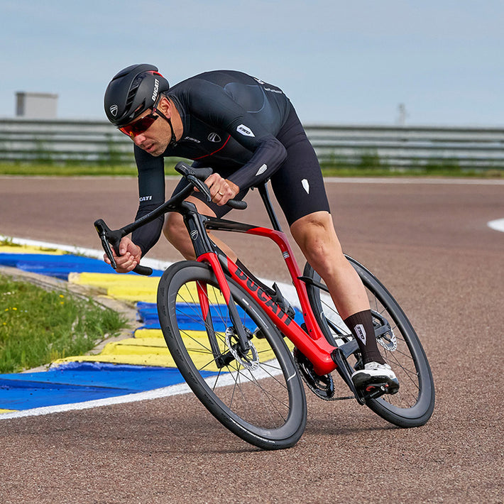 A cyclist in a black cycling kit leans forward on a red and black road bike on an outdoor track with blue and yellow curbs